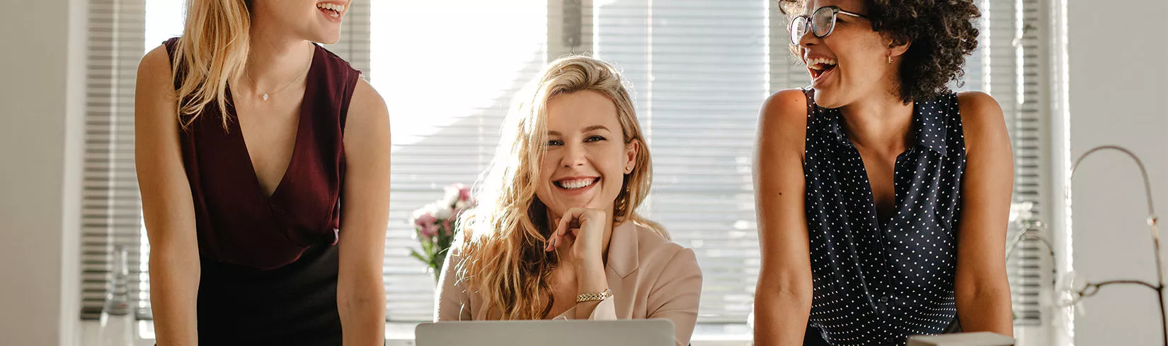 Drei junge Frauen stehen/sitzen im Büro und lachen herzhaft